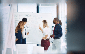 A diverse team of business professionals collaborates around a whiteboard covered in strategic planning notes during a marketing planning session.