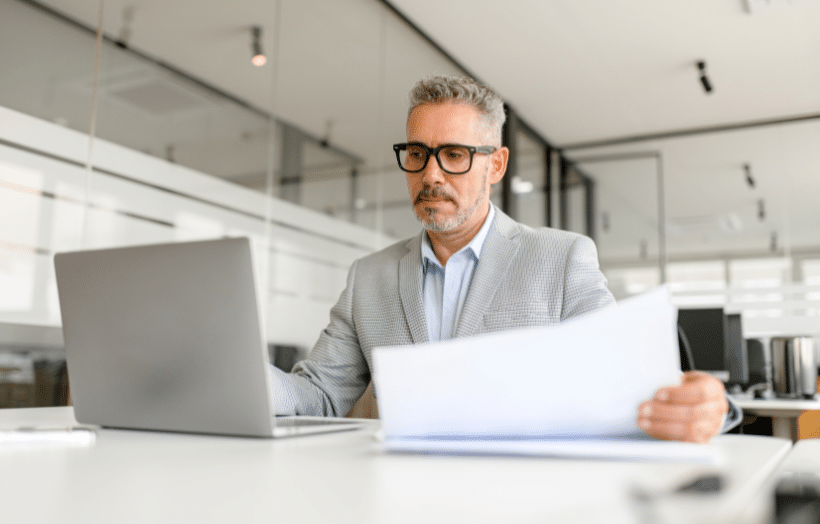 Senior business professional reviewing documents at laptop in modern office