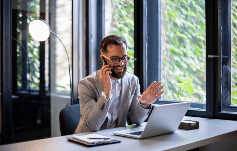 Businessman smiling on phone call at desk with laptop in modern office