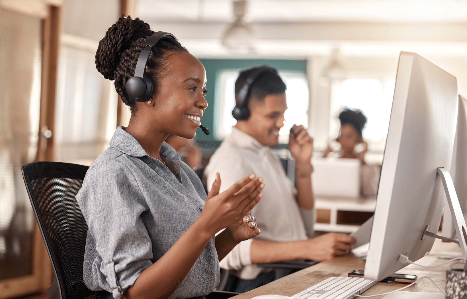 Call center associate wearing a headset and smiling while on a call, with teammates working in the background