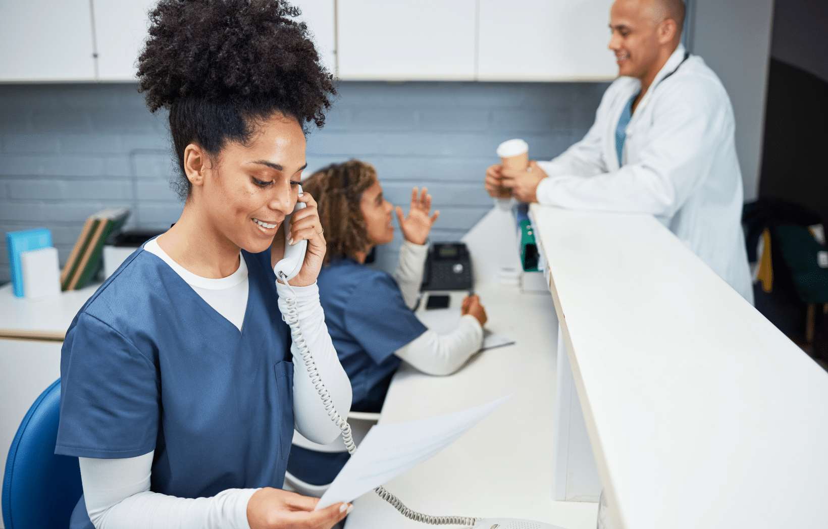 Smiling healthcare worker in blue scrubs talking on the phone at a clinic front desk